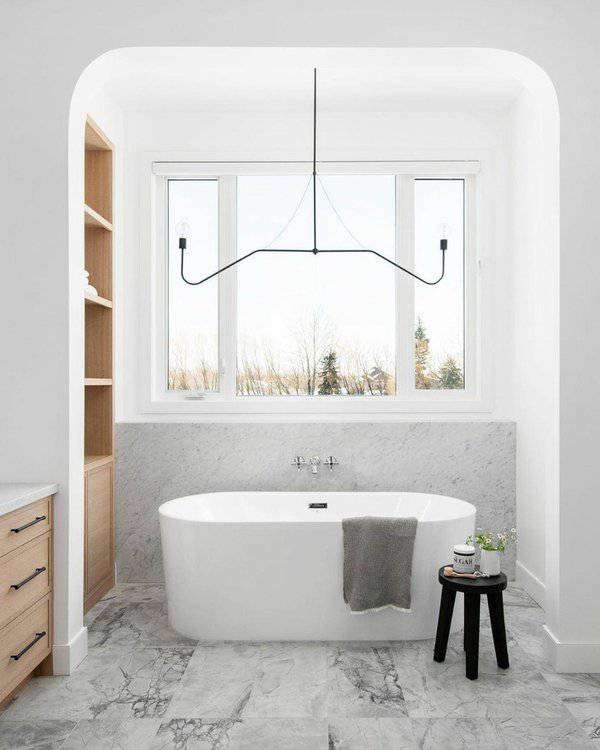 Slim wire chandelier above a white freestanding tub with marble flooring and backsplash in a minimalist, light-filled bathroom.