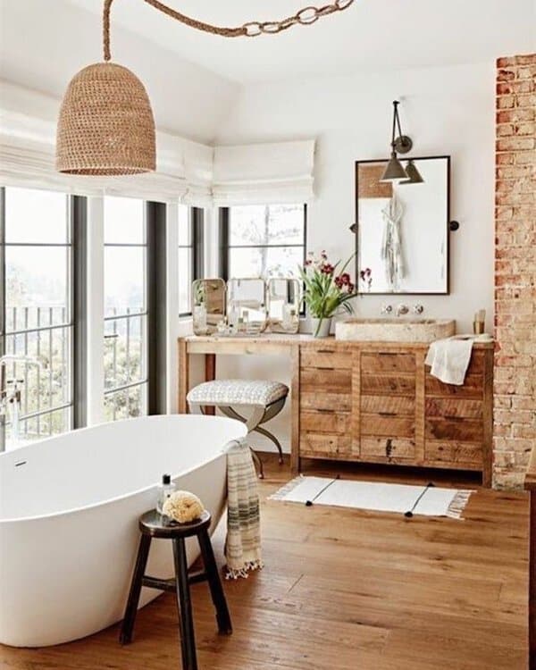 Woven pendant above a white freestanding tub, wood vanity, exposed brick, and large windows in a relaxed, rustic-chic bathroom.