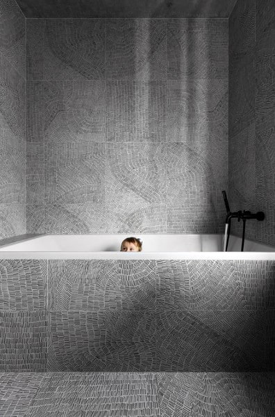 Bathroom with intricate patterned gray tiles and a built-in bathtub with black faucet.