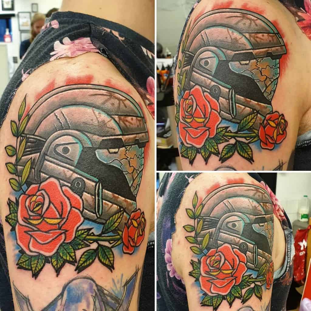 Stunning Halo tattoo of a cracked Spartan helmet surrounded by vibrant red roses and green leaves, blending sci-fi and nature on the upper arm.