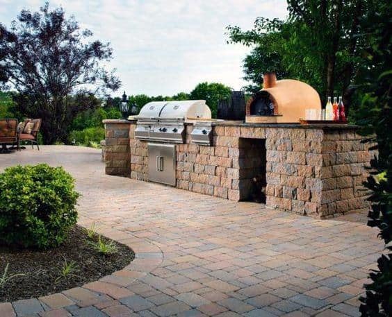 Outdoor kitchen with grill, pizza oven, and stone countertops on a patio surrounded by lush greenery and trees