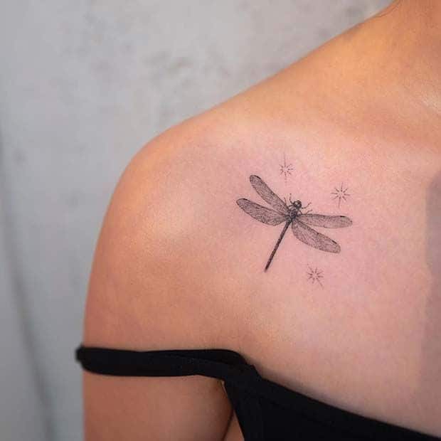 Delicate black ink dragonfly tattoo on the shoulder with subtle star accents, creating a minimalist and elegant design.