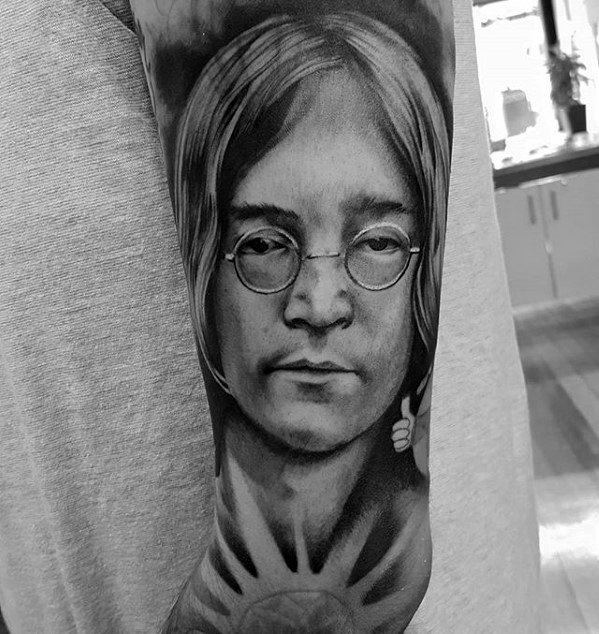 Hyper-realistic black and gray tattoo of John Lennon wearing round glasses, with detailed facial shading and a subtle peace sign below.