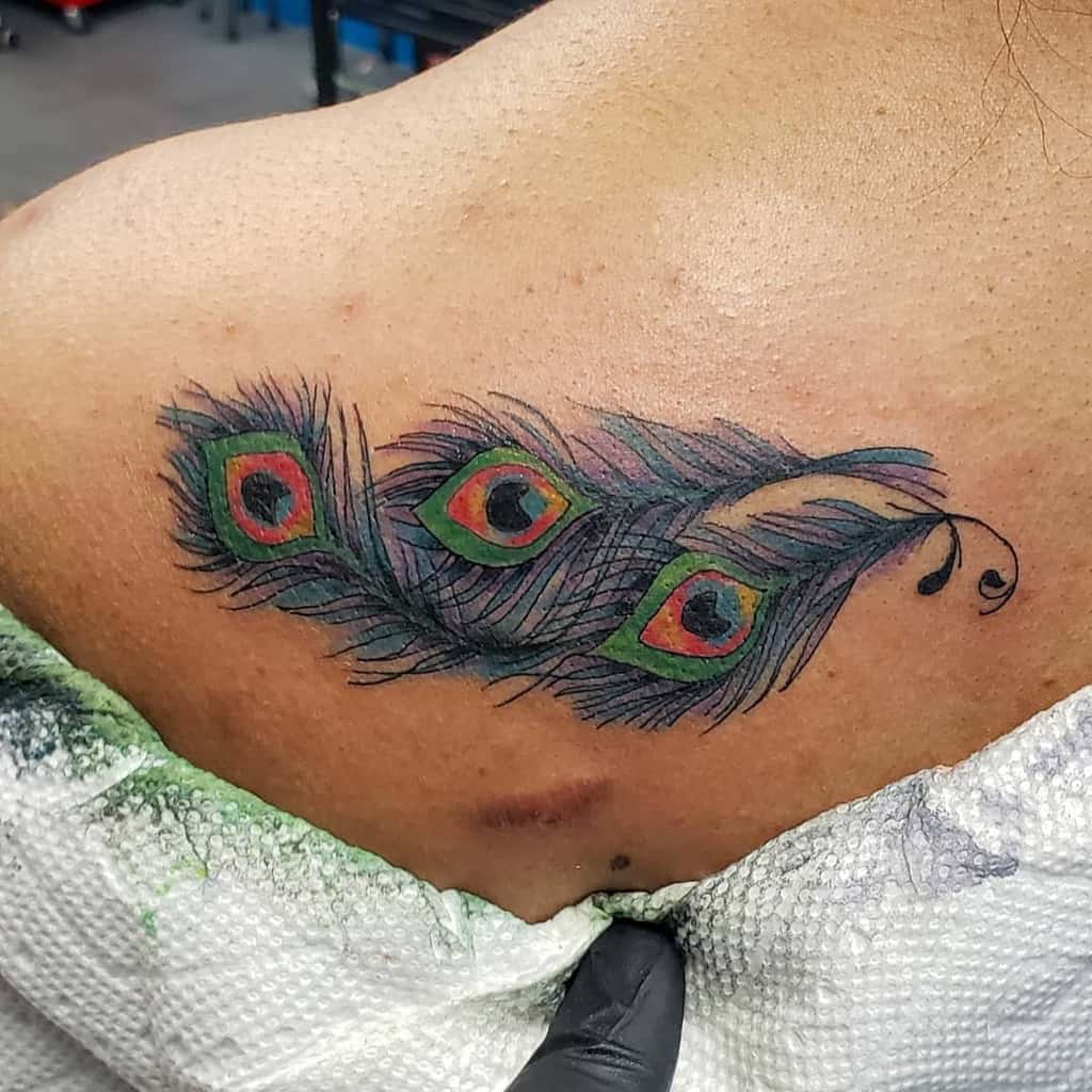 Colorful peacock feather tattoo on the upper back, featuring three eye patterns with green, red, and blue hues, along with fine shading.