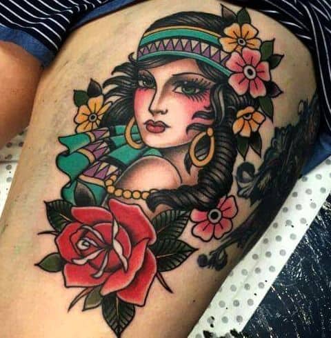 Vibrant tattoo of a gypsy woman with a patterned headband, gold earrings, and colorful flowers, featuring bold shading on the thigh.
