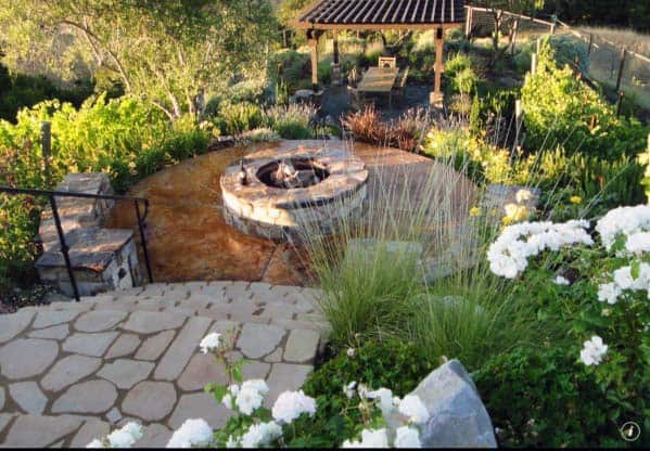 Stone patio with fire pit, surrounded by lush greenery, white flowers, and a pergola in the background