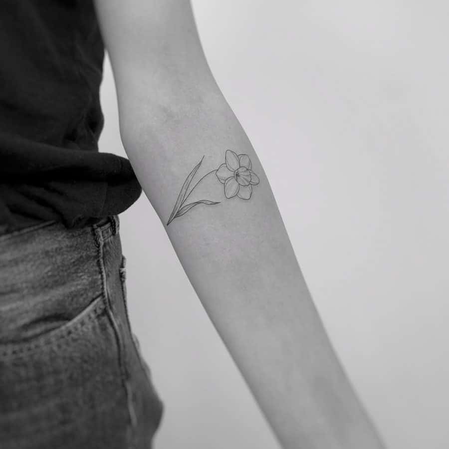A delicate black-and-white tattoo of a single daffodil with a slender stem and leaves, offering a minimalist yet elegant design.