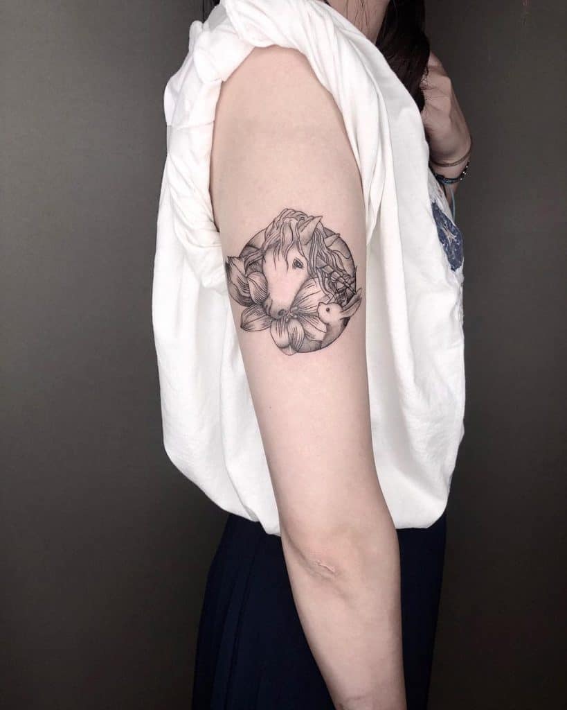 Delicate black ink tattoo of a unicorn’s head surrounded by flowers and a butterfly, creating a soft and elegant design on the upper arm.