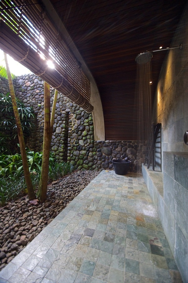 Luxurious outdoor shower with stone walls, bamboo accents, and tiled flooring surrounded by greenery.