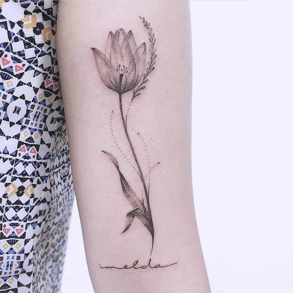 A delicate black and grey tulip tattoo on the forearm, featuring fine shading, botanical details, and the name "Imelda" in cursive script.
