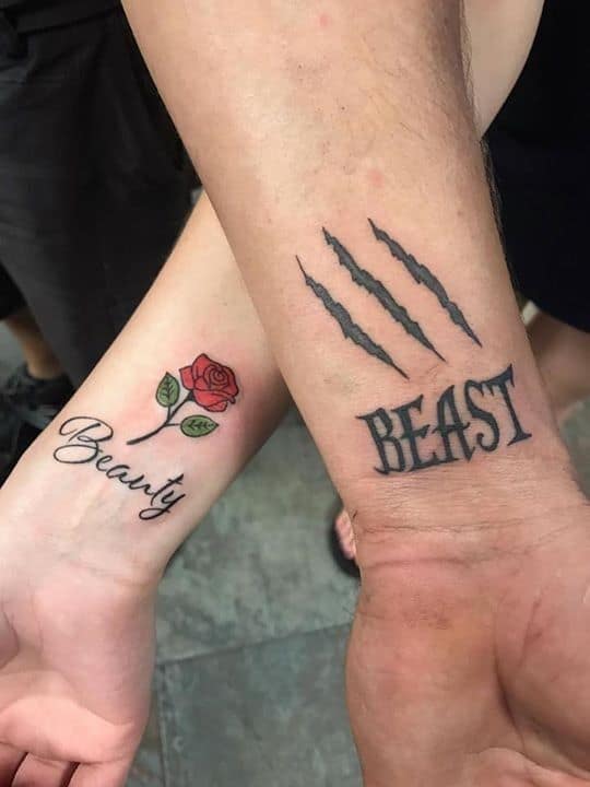 Matching couple tattoos with 'Beauty' and a red rose on one wrist, and 'Beast' with claw marks on the other, symbolizing their connection.