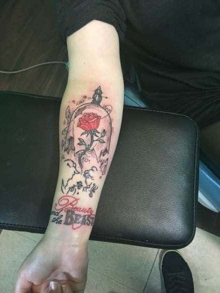Forearm tattoo of the enchanted rose inside a glass dome, surrounded by swirling details, with 'Beauty and the Beast' written in red script.