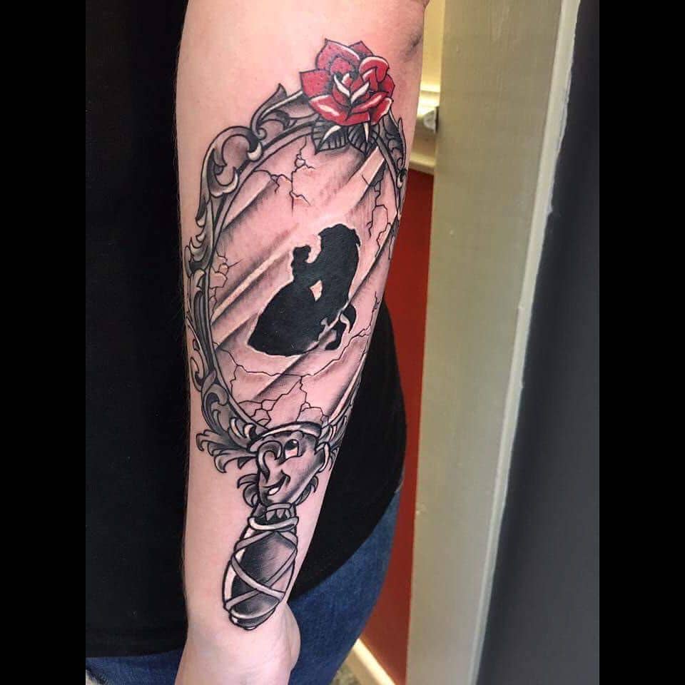 Ornate mirror tattoo with a cracked glass effect, featuring Belle and the Beast’s silhouette, accented by a red rose at the top.