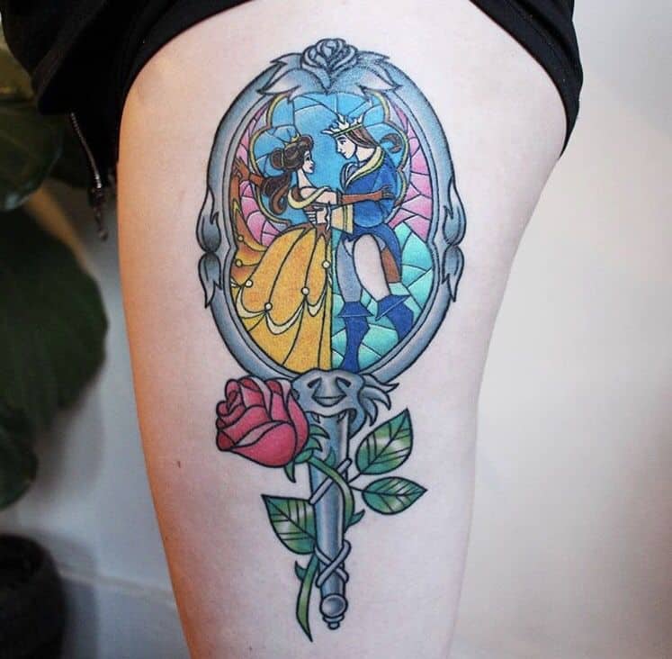 Stained glass-style tattoo of Belle and the Beast dancing, framed within an ornate hand mirror with a red rose and vine details.