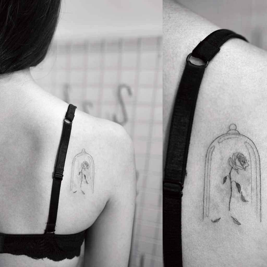 Minimalist fine-line tattoo of the enchanted rose inside a glass dome, placed on the upper back with delicate shading and falling petals.