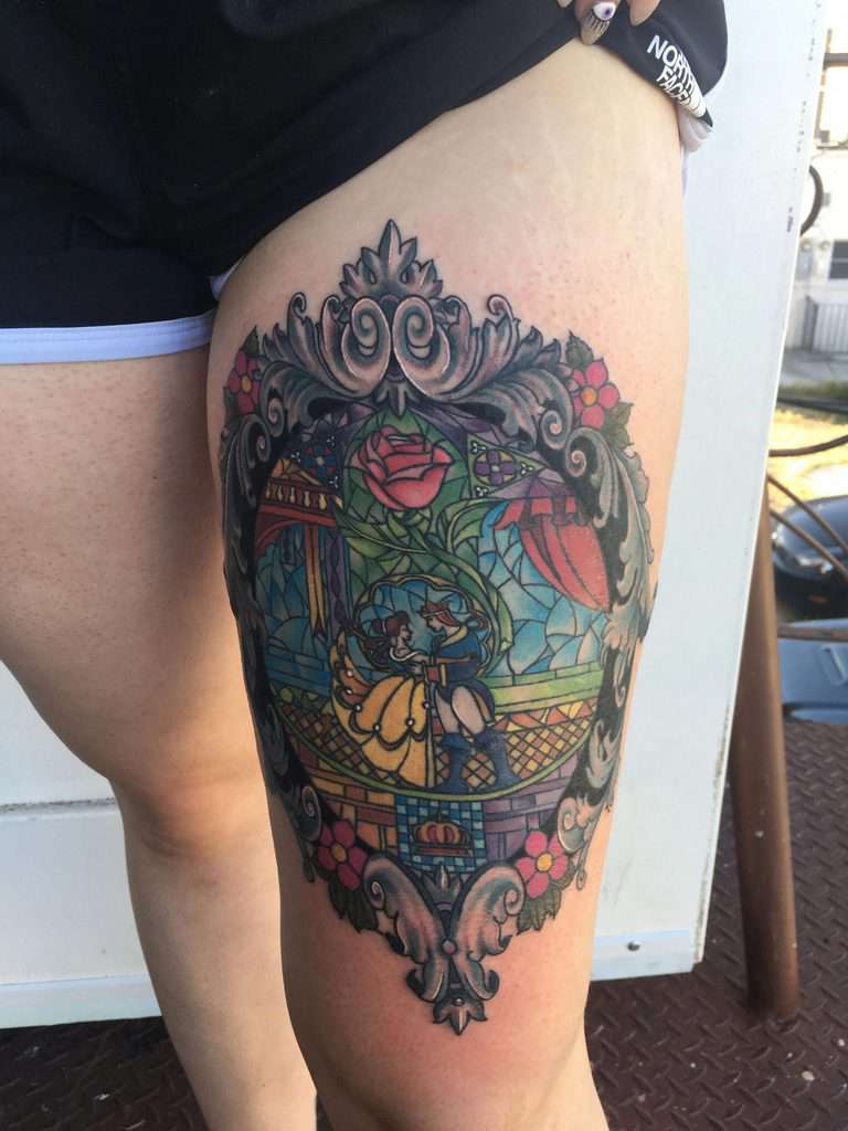 Large stained glass-style thigh tattoo of Belle and the Beast dancing, framed in an ornate design with intricate detailing and rich colors.