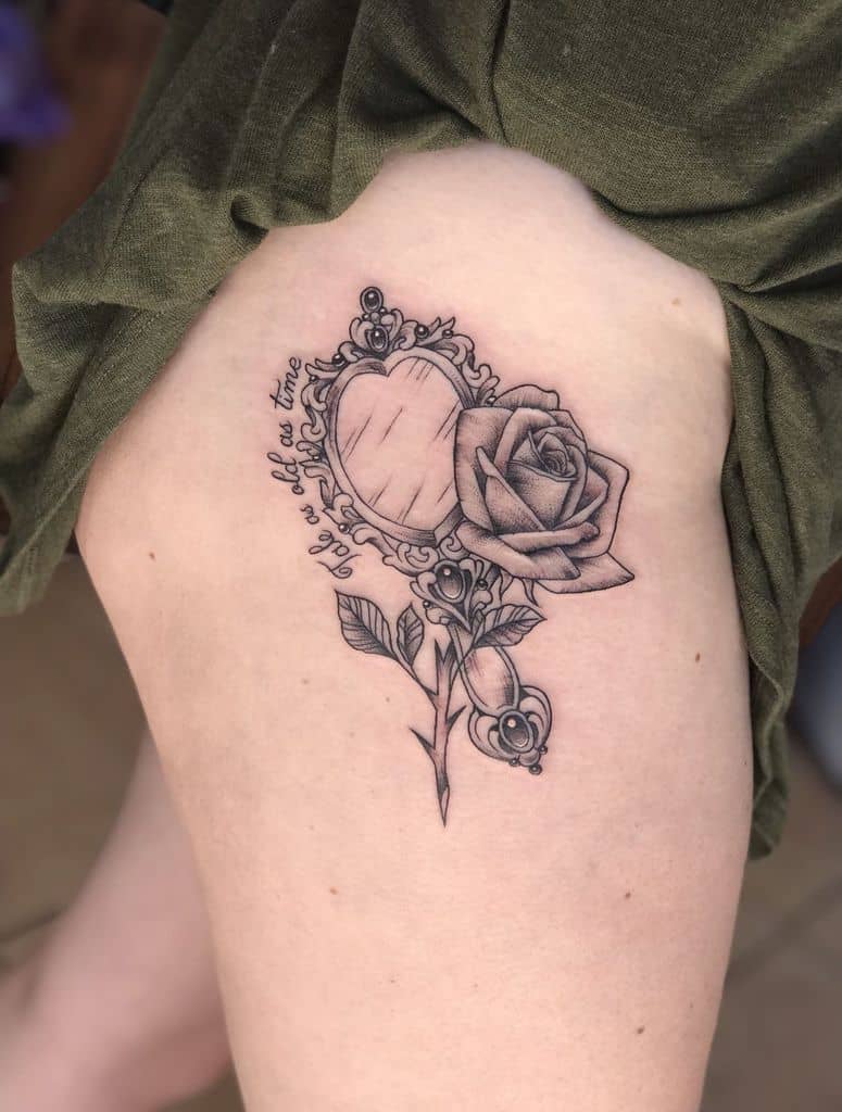 Elegant black and grey tattoo featuring an ornate mirror, a detailed rose, and the phrase 'Tale as old as time' in cursive on the thigh.