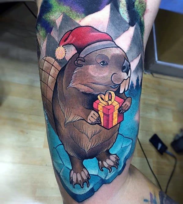 Festive beaver tattoo wearing a Santa hat and holding a gift box while standing on ice, with colorful mountains in the background.