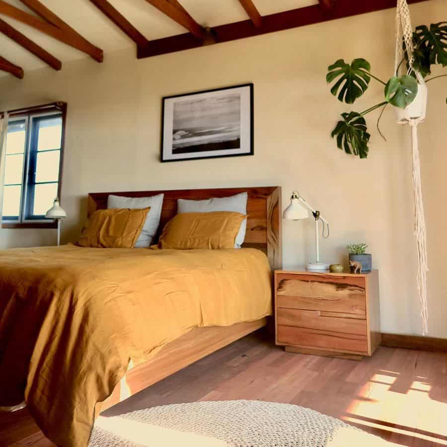 Cozy bedroom with a wooden bed, yellow bedding, a nightstand, and a hanging plant under wooden ceiling beams