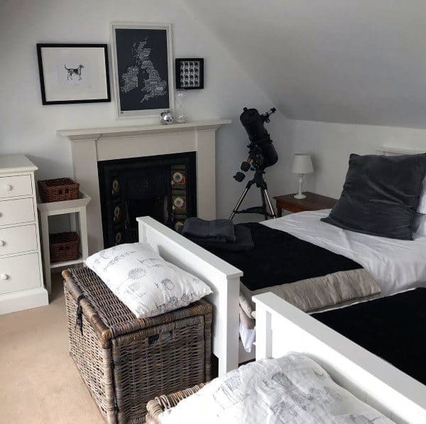 Cozy attic bedroom with twin beds, telescope, and framed art above a fireplace, white walls and wicker accents