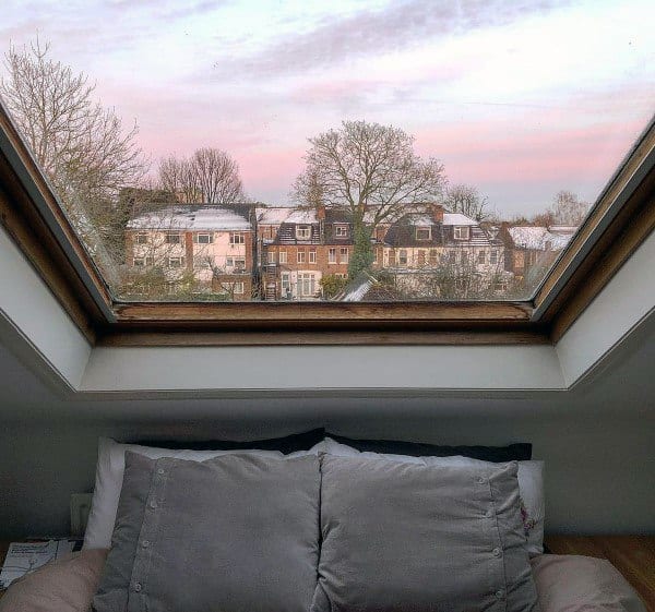 View from an attic skylight showing a cozy bed and snow-dusted rooftops at sunrise or sunset
