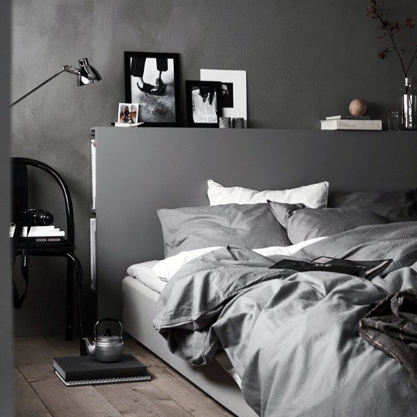 A cozy bedroom with grey bedding, a lamp, books, photos on a headboard shelf, and a chair with a phone