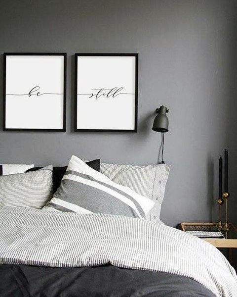 A minimalist bedroom with grey walls, striped bedding, framed "be still" prints, and a wall lamp