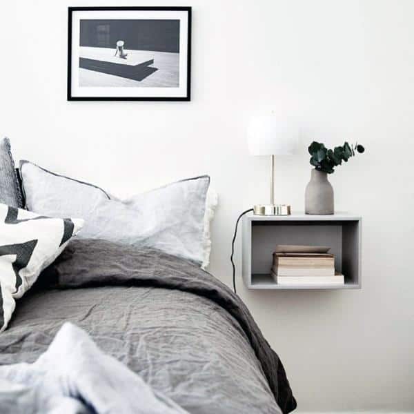 Minimalistic bedroom with grey bedding, framed art, and a small shelf holding a lamp, books, and a vase on the wall