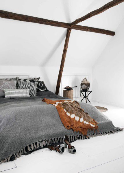 Cozy attic bedroom with exposed beams, patterned pillows, a gray blanket, and heeled shoes scattered on the floor