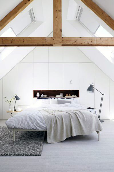 Minimalist white bedroom with exposed wooden beams, skylights, a neatly made bed with soft linens, and a floor lamp