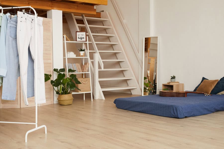 Minimalist room with blue mattress on the floor, clothes rack, potted plants, large mirror, and wooden staircase leading to a loft