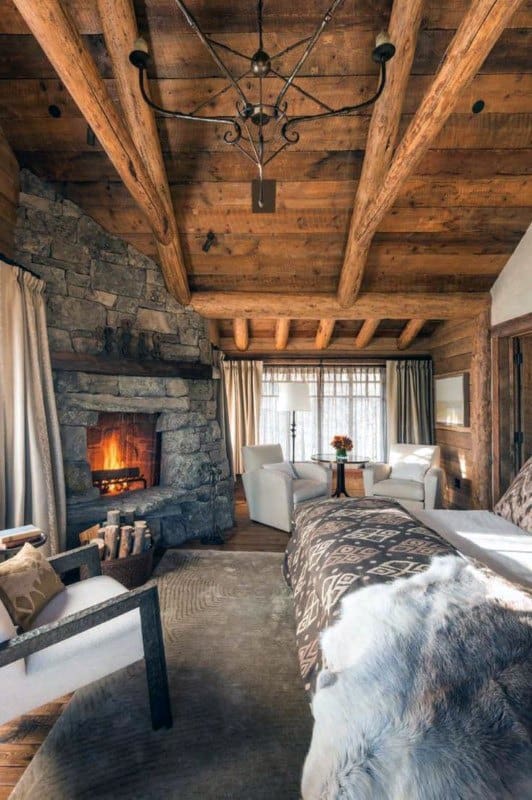 Rustic bedroom with wooden beams, stone fireplace, patterned bedding, and cozy seating area.