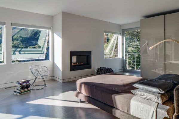 Modern bedroom with large windows, recessed fireplace, and concrete flooring with flush baseboards.