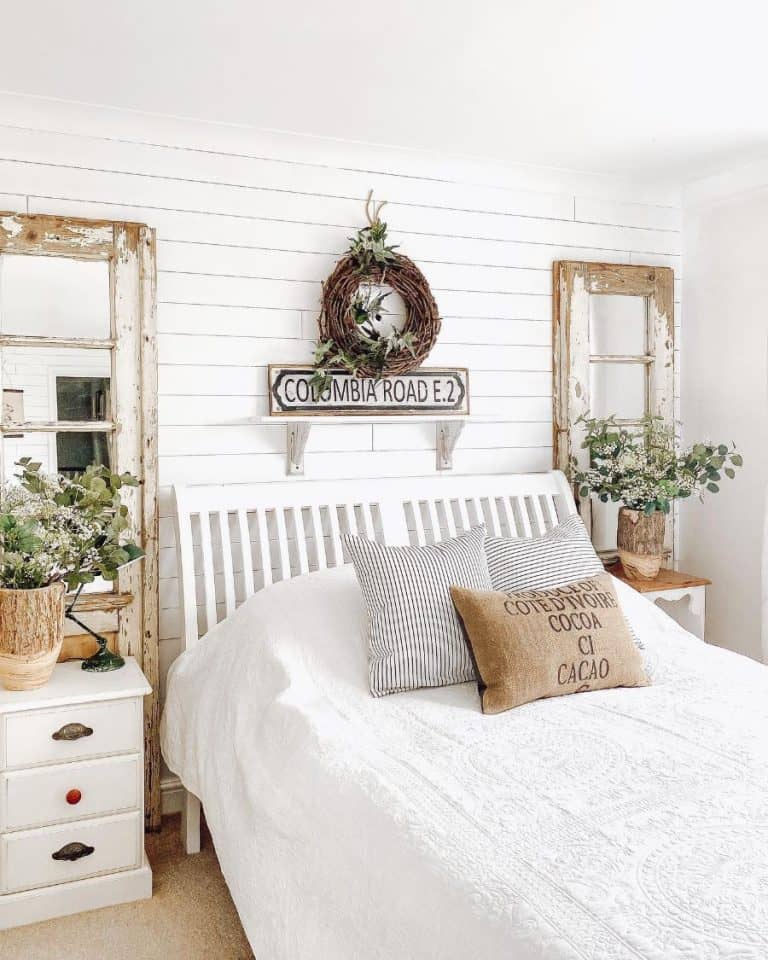 Farmhouse-style bright bedroom with a white bed, rustic mirrors, and potted plants; a wreath and sign hang above the bed