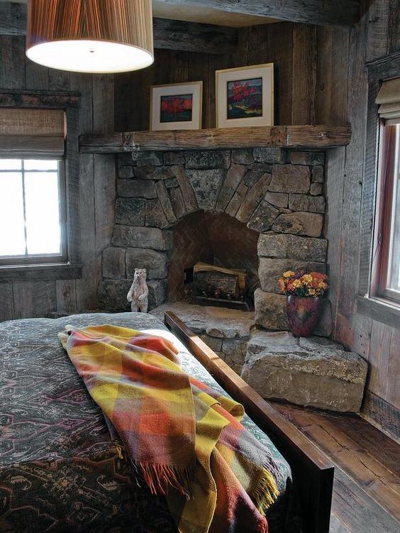 Rustic bedroom with stone corner fireplace, wooden mantel, warm pendant lighting, and colorful blanket.