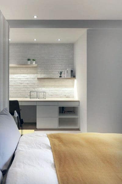 Minimalist bedroom with a beige blanket, white brick wall, and shelves with small plants in the background