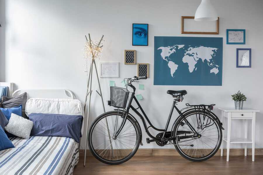 A black bicycle leans against a white wall in a modern room with a blue world map, framed pictures, a sofa with blue cushions, and a small white table with a plant