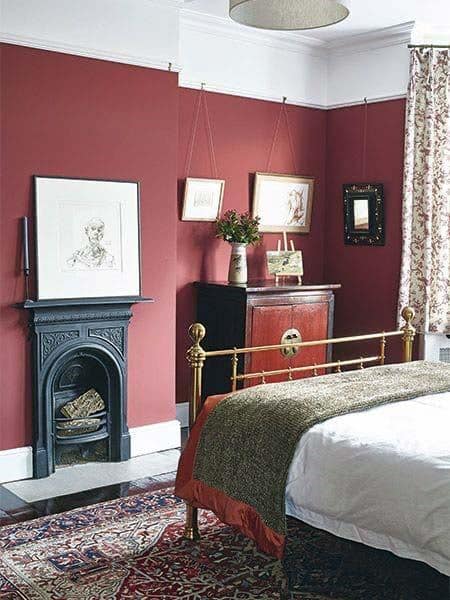 Cozy bedroom with a brass bed, bold red walls, a small fireplace, framed art, and floral curtains