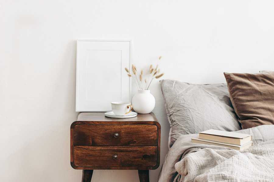 Wooden bedside table with a white vase, cup, and books in a cozy bedroom setting.