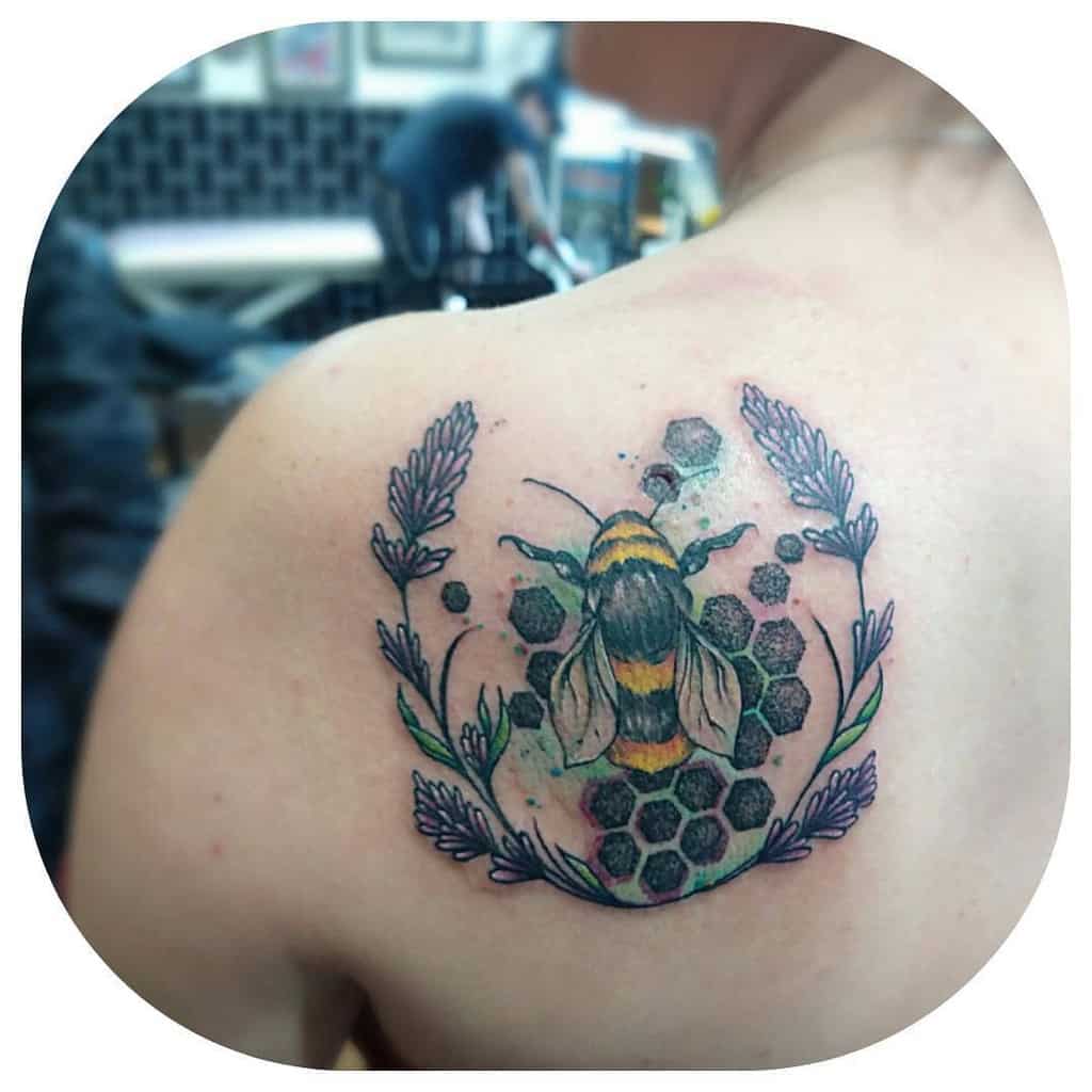 A tattoo of a bumblebee hovering over honeycomb with lavender accents, surrounded by soft watercolor splashes, located on the upper back.