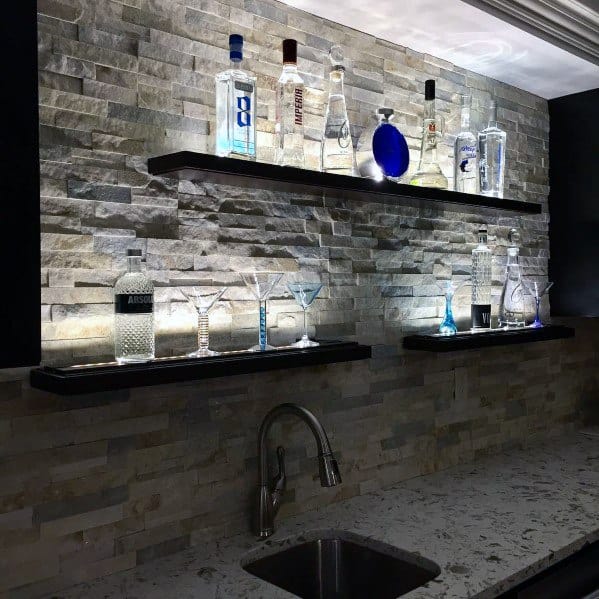 Home bar with beige stacked stone backsplash, floating shelves, LED lighting, and a stylish display of bottles and glasses