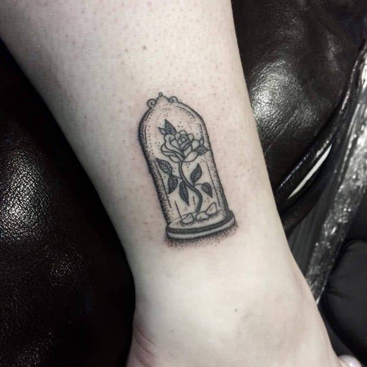 Black and grey dotwork tattoo of the enchanted rose inside a glass dome, placed on the ankle with fine-line detailing.