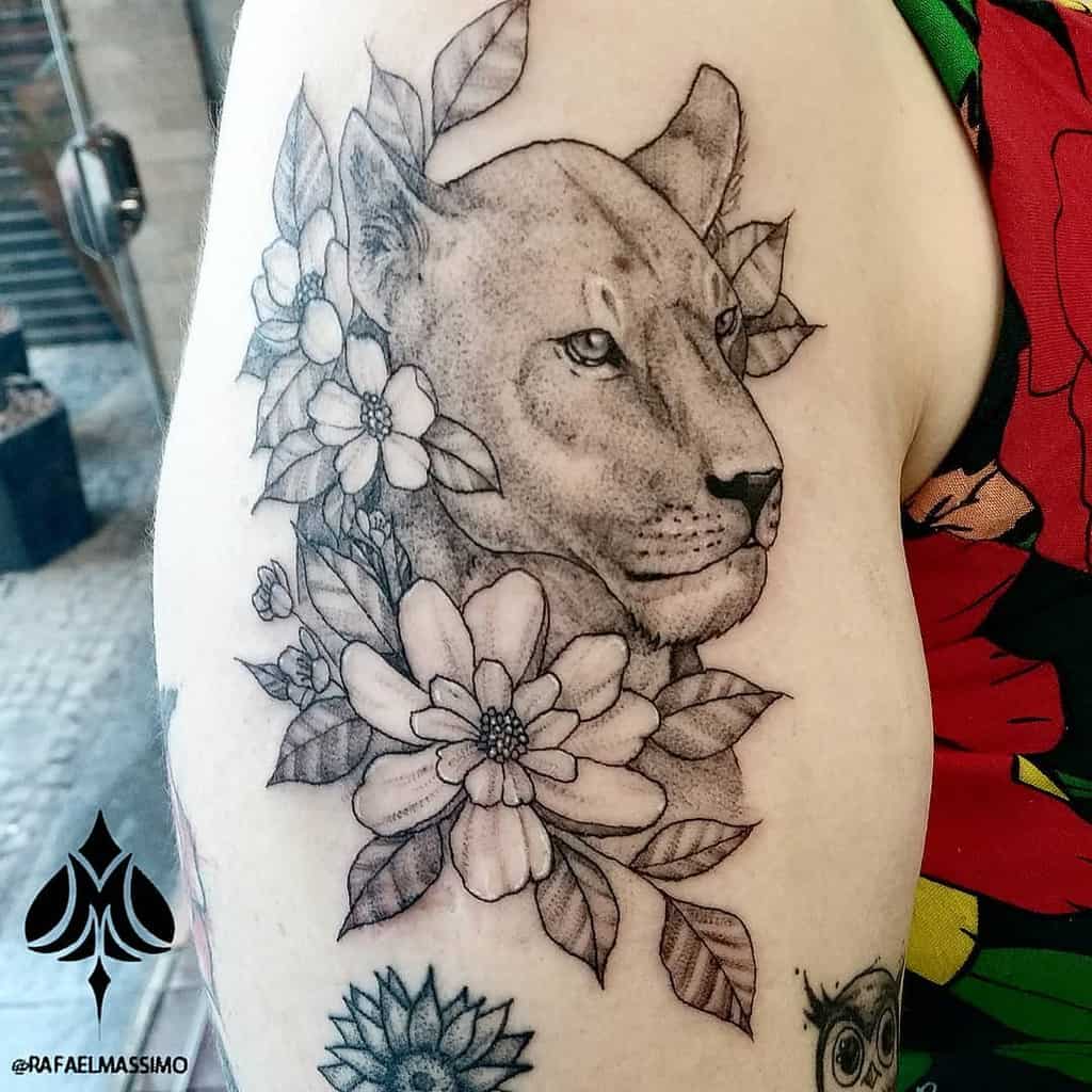 Realistic lioness tattoo on the upper arm, surrounded by detailed flowers and leaves, blending strength and femininity with fine shading.
