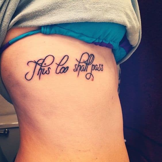 A graceful "This too shall pass" tattoo in cursive script, placed on the ribcage, symbolizing resilience and hope.
