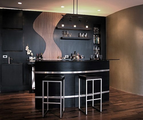 Modern bar with black counter, stools, and shelves for bottles and glasses against a wood and black wall