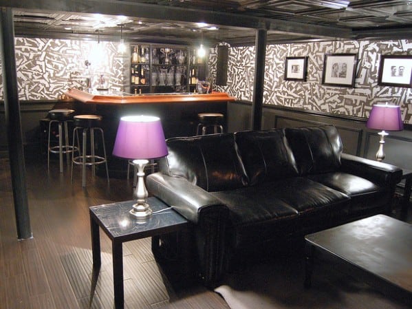 Chic basement man cave with a black leather sofa, corner bar, barstools, purple lamps, and patterned wallpaper