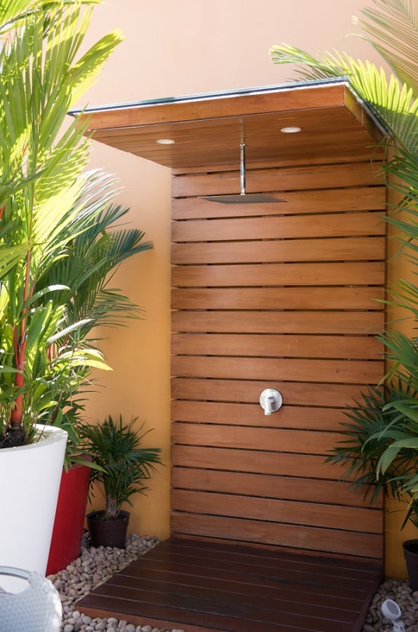 Modern outdoor shower with wooden slat wall, pebble flooring, and potted tropical plants.