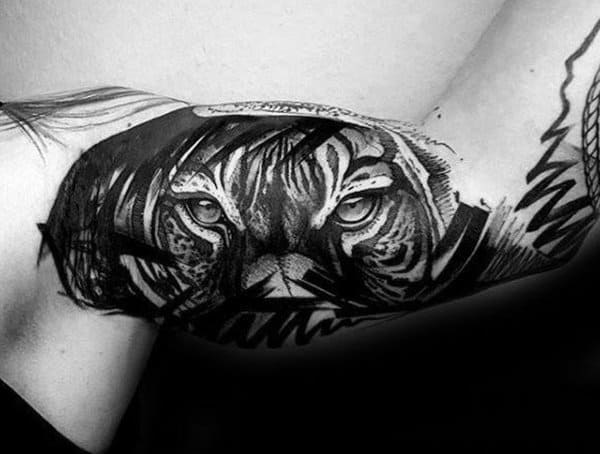 Inner bicep tattoo for men showcasing a fierce tiger face in black and grey realism with artistic brushstroke effects.