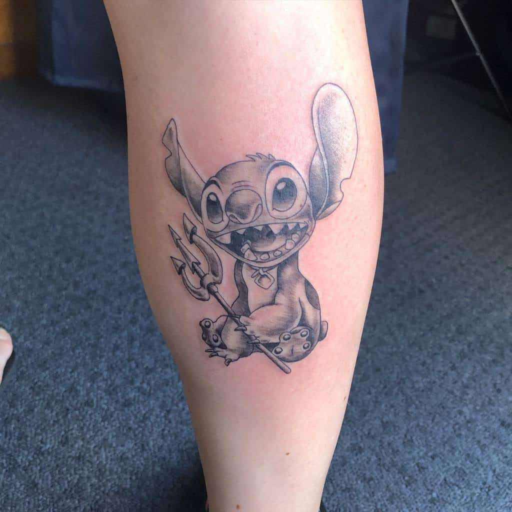 Black and gray tattoo of Stitch from Lilo & Stitch holding a trident, with a playful grin, on the calf.