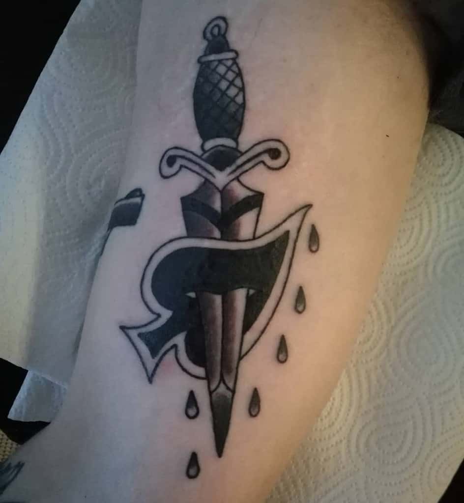 A black-and-white tattoo of a dagger piercing through an Ace of Spades symbol, with tears dripping down around the design